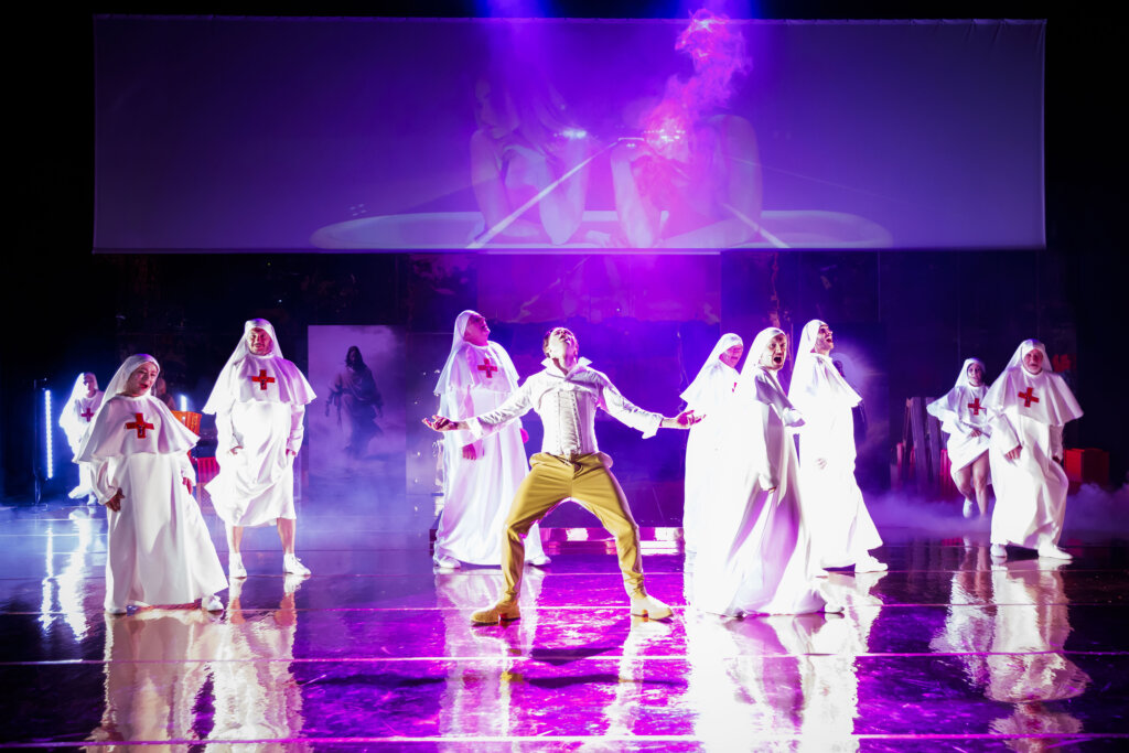 Młody mężczyzna w koszuli, gorsecie i złotych spodniach tańczy na środku sceny. Ma rozłożone ręce i twarz skierowaną ku górze. Wokół niego tańczy 9 aktorów i aktorek w białych kostiumach zakonnic.